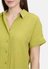 Korte mouwen collomshirt in lichtgroen, gemaakt van lichte stof. Heeft een knoopsluiting en opgerolde mouwdetails.