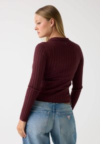 Burgunderfarbener, gerippter Langarmoberteil mit einem runden Kragen und einer einzelnen Knopfdetails auf der Rückseite, kombiniert mit hellblauen, hoch taillierten Jeans.