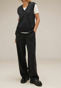 Black knitted sleeveless vest with a V-neck over a white T-shirt, paired with loose-fitting black trousers and chunky black shoes.