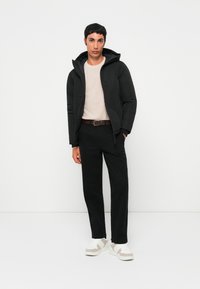 Jonge man staat gekleed in een zwarte hoodie, beige sweater, zwarte broek, bruine riem en witte sneakers voor een effen witte achtergrond.