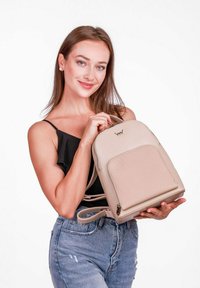 Béžový batoh z hladkého syntetického materiálu, zaoblený tvar, přední kapsa a detail s logem, držený osobou v černém tričku.