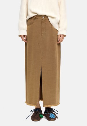 Osoba nosząca beżowy sweter, brązową midi spódnicę z przednim rozcięciem i frędzlami na dole oraz ciemne trampki z niebiesko-zielonymi ozdobnymi sznurówkami.