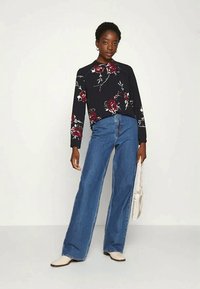 Blusa preta com estampado floral e mangas compridas, combinada com jeans azuis de perna larga e cintura alta. A modelo segura uma bolsa clara.