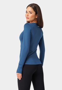 Zilās krāsas garām piedurknēm sporta krekls, ar pieguļošu dizainu, gludu tekstūru un pirkstu caurumiem. Kombinēts ar ribotām melnām legingu.