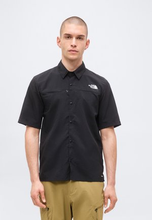 Mand iført en sort kortærmet skjorte med en lille hvid logo på brystet og khaki cargobukser, stående mod en ensfarvet baggrund.