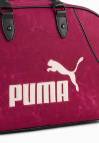 Bordowa torba z płótna z czarnymi skórzanymi akcentami, z dużym białym logo "PUMA" oraz sylwetką skaczącego kota. Gładka tekstura, wyraźny kształt.