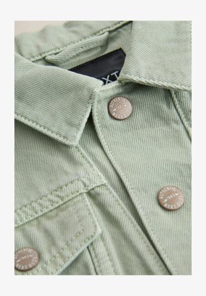 Chemise en denim vert clair avec col pointu, fermeture par boutons et éléments décoratifs en bronze gravés "Authentic 1982".