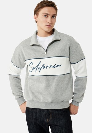 Young man wearing a grey quarter-zip sweatshirt with white panels and "California" written across the chest, paired with dark jeans.