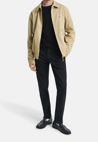 Veste beige avec un col, pull noir en dessous, pantalon noir et chaussures noires cirées. La veste comporte des poches latérales et un détail logo.