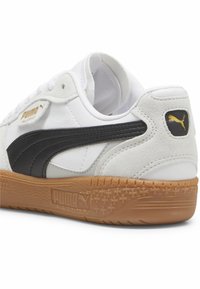 Bílé tenisky s černým koženým pruhem, zaměněnými akcenty a gumovou podrážkou. Na vnější straně se nachází zlatě embosované logo.