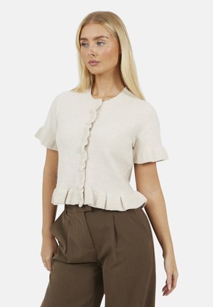 Blonde vrouw die een crèmekleurige korte mouw gerimpelde cardigan en hoge taille bruine geplooide broek draagt, poserend tegen een effen achtergrond.