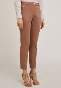 Pantaloni marroni aderenti con una texture liscia, caratterizzati da un dettaglio con bottone sul lato e abbinati a sandali con tacco alto e cinturini.