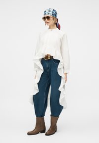 Mujer con blusa blanca de volantes de gran tamaño, vaqueros azules de talle alto, botas marrones, gafas de sol y pañuelo azul con estampado, de pie frente a un fondo blanco.