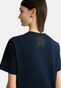 T-shirt blu navy a maniche corte con un'etichetta logo nera testurizzata sul retro. Materiale in cotone morbido, con una vestibilità rilassata e un design minimal.