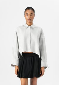 Chemise courte à manches longues, en gris clair avec des rayures blanches verticales et un col à boutons, associée à une jupe noire avec une ceinture élastique.