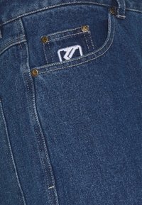 Jeans de denim azul con un bolsillo trasero que lleva un parche de logo blanco y remaches de tono dorado. La tela tiene un tejido texturizado y un cosido clásico.