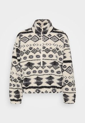 Fleece-pullover med høy krage, med hvit bakgrunn og svarte geometriske mønstre, inkludert diamanter og stiliserte former.
