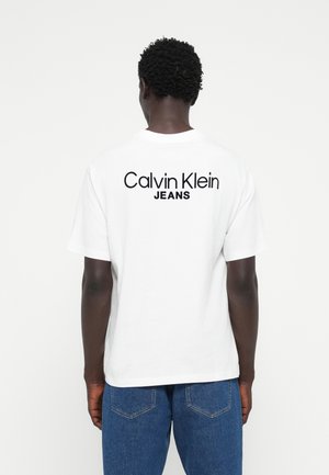 White cotton t-shirt featuring a black "Calvin Klein JEANS" logo printed on the back. Short sleeves and a crew neckline.