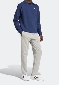 Sudadera azul marino con cuello acanalado y logo blanco, combinada con pantalones de chándal gris claro. El modelo lleva zapatillas blancas con detalles negros.