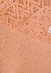 Perzikkleurige stof met een gestructureerd breipatroon, voorzien van verfijnde kantdetails met zigzag- en bloemmotieven in het bovenste gedeelte.