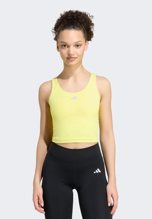 Donna con capelli ricci indossa un top corto giallo senza maniche e leggings neri a vita alta, in piedi davanti a uno sfondo chiaro e semplice.