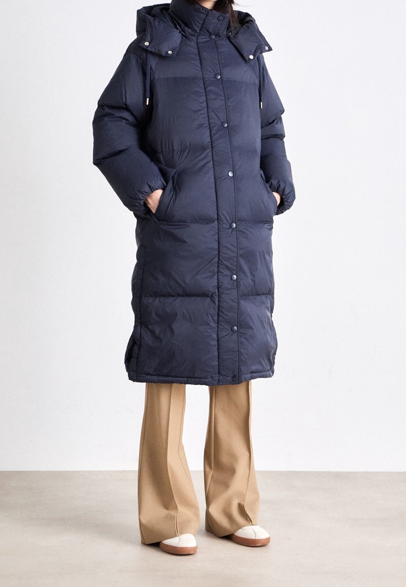 Marineblaue Pufferjacke mit knielangem Design, hohem Kragen und abnehmbarer Kapuze. Verfügt über große Taschen und eine Knopfleiste vorne.