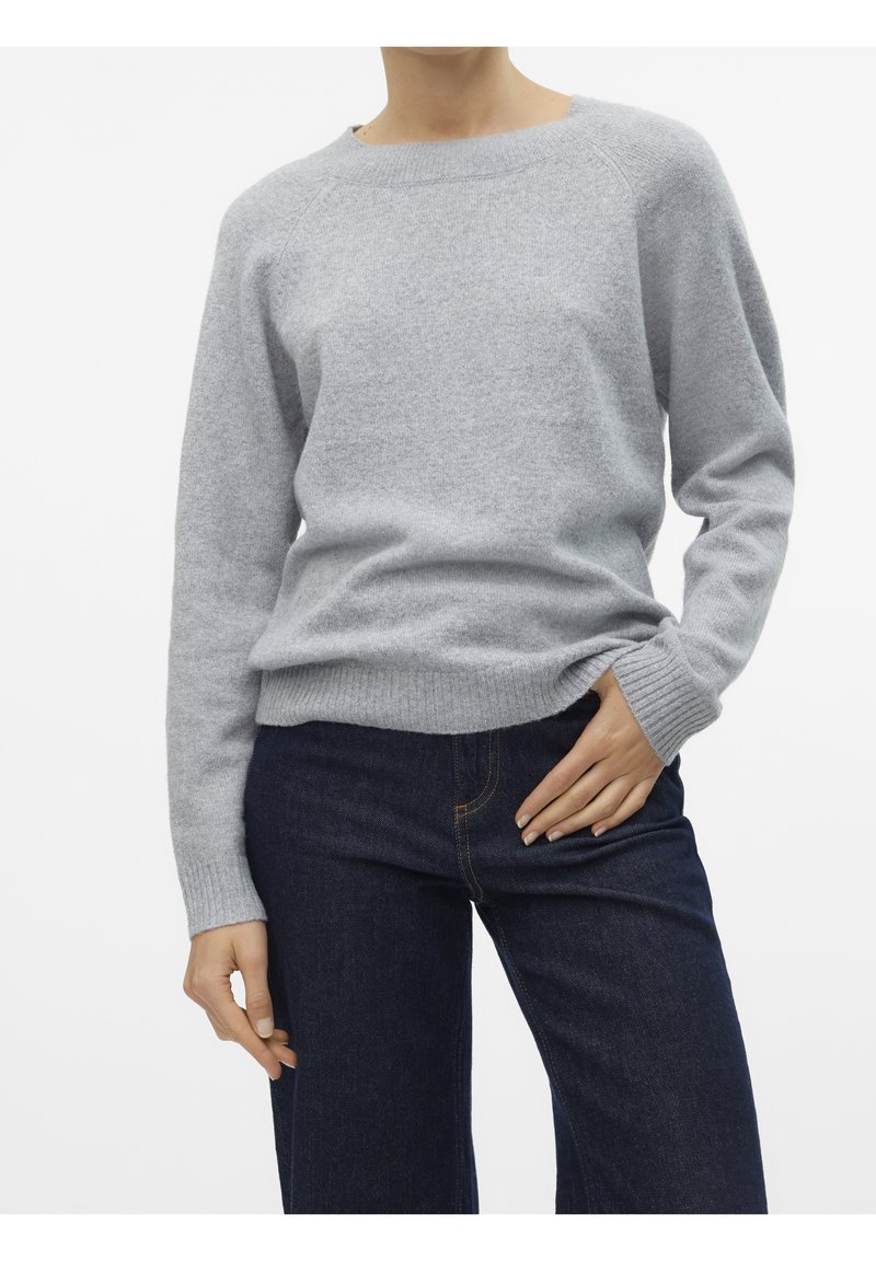 Personne portant un pull gris clair à manches longues et col rond, ainsi qu'un jean bleu foncé, avec une main dans la poche et l'autre détendue le long du corps.