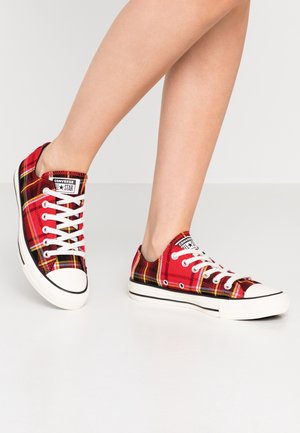 Personne portant des baskets Converse basses à carreaux rouges avec des lacets blancs et des semelles en caoutchouc, debout sur un sol clair.
