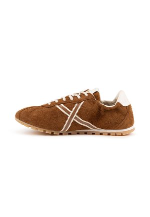 Zapatilla deportiva de ante marrón con cordones blancos, lengüeta trasera blanca y franja blanca en forma de X en el lateral, mostrada desde el lado exterior sobre un fondo blanco.