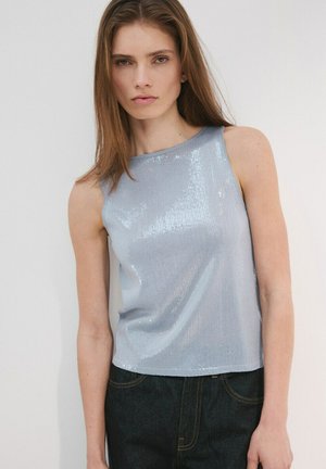 Mujer joven con cabello castaño lacio que lleva una blusa sin mangas azul claro con lentejuelas y jeans oscuros, de pie contra un fondo liso y claro.