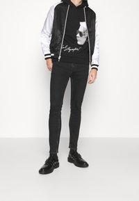Chaqueta bomber en blanco y negro con acabado satinado, con capucha y puños rayados, llevada sobre una sudadera gráfica negra y jeans ajustados.