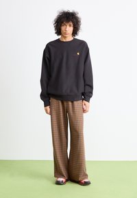 Zwarte sweatshirt met een klein logo, bruine en beige geruite wijde broek, en zwarte schoenen met rode accenten. Plat materiaal, relaxte pasvorm.