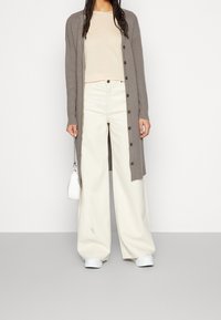 Długa szara kardiganka, beżowa dopasowana bluzka, białe szerokie spodnie, białe sneakersy oraz mała biała torebka na łańcuszku. Kardiganka ma guziki.