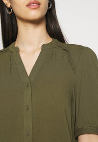 Blusa leggera verde oliva, con chiusura a bottoni sul davanti, dettagli a scallop sulle maniche e scollatura rotonda con pieghe sottili.