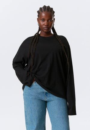 Maglietta nera a maniche lunghe con vestibilità rilassata, scollo tondo e dettaglio annodato sul lato, abbinata a jeans a gamba dritta di colore azzurro chiaro.