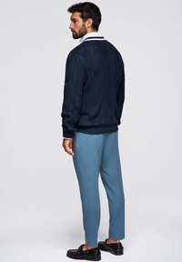 Granatowy sweter dzianinowy z geometrycznymi wzorami, zestawiony z jasnoniebieskimi spodniami w kant oraz czarnymi mokasynami. Biała koszula z kołnierzykiem widoczna na szyi.