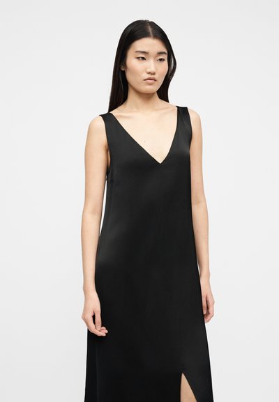 Νεαρή γυναίκα με ίσια μαύρα μαλλιά, ντυμένη με αμάνικο μαύρο φόρεμα με βαθύ V ντεκολτέ και πλευρική σχισμή, στέκεται μπροστά σε απλό λευκό φόντο.