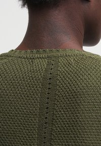 Oliivivihreä neulottu neulepusero, jossa on teksturoitu kuvio, ribbireunus kauluksessa ja pystysuora rivi pieniä aukkoja takasaumassa.