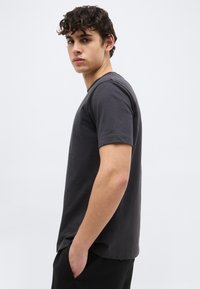 Νεαρός άνδρας με σγουρά μαλλιά στέκεται σε προφίλ, φοράει ένα απλό σκούρο γκρι κοντομάνικο μπλουζάκι και μαύρο παντελόνι μπροστά σε ανοιχτό φόντο.