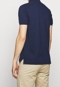 Marinblå piké med korta ärmar, platt krage, sidoslitsar, gjord av glatt tyg och med en diskret textur. Bärs tillsammans med beige byxor.