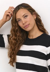 Pull rayé avec de larges bandes horizontales noires et blanches, col rond et un tissu en tricot doux et texturé. Modèle avec des cheveux ondulés.