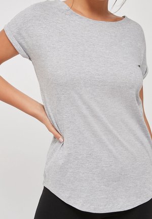 Frau trägt ein hellgraues kurzärmeliges T-Shirt mit hochgekrempelten Ärmeln, eine Hand auf der Hüfte ruhend, vor einem schlichten hellen Hintergrund.