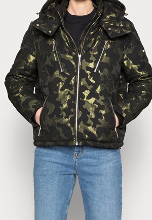 Mężczyzna ubrany w czarno-złotą kurtkę z kapturem o wzorze moro z srebrnymi zamkami, ręce w kieszeniach, w połączeniu z jasnoniebieskimi dżinsami.