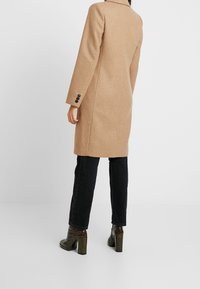 Cappotto in misto camel con maniche lunghe, spacco sul retro e dettagli con bottoni neri, abbinato a jeans neri aderenti e stivaletti neri con tacco dal tessuto texturizzato.
