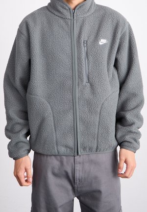 Personne portant une veste polaire grise zippée avec une poche verticale sur la poitrine et le logo blanc Nike sur le côté gauche.