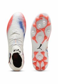 Weiße Fußballschuhe mit einer strukturierten Oberfläche, die rot-blau verlaufende Muster aufweisen. Helle orange Stollensohle mit einem schwarzen Logo.