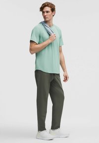 Ung man i ljusgrön T-shirt, mörkgröna byxor, vita sneakers, som håller en ljusgrå sweatshirt över axeln, stående mot en enfärgad bakgrund.