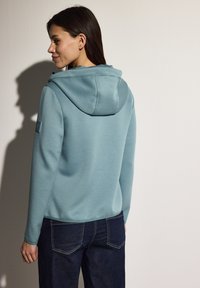 Felpa blu chiaro con cappuccio e coulisse, dal tessuto morbido, abbinata a jeans in denim scuro con dettagli di cucitura visibili.