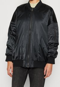Veste bomber en satin noir avec poignets et col côtelés. Comprend des poches latérales et une fermeture éclair. Texture lisse, coupe décontractée.