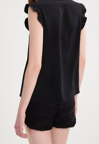 Schwarzes Top mit Rüsche an den Ärmeln, kombiniert mit hoch taillierten schwarzen Shorts, die horizontale Falten und Fransenverzierungen am Saum aufweisen.